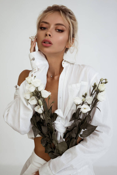 beautiful blonde model posing in white clothes. woman with updo hairstyle. natural make-up look. glamour lady holding flowers - Fotó, kép