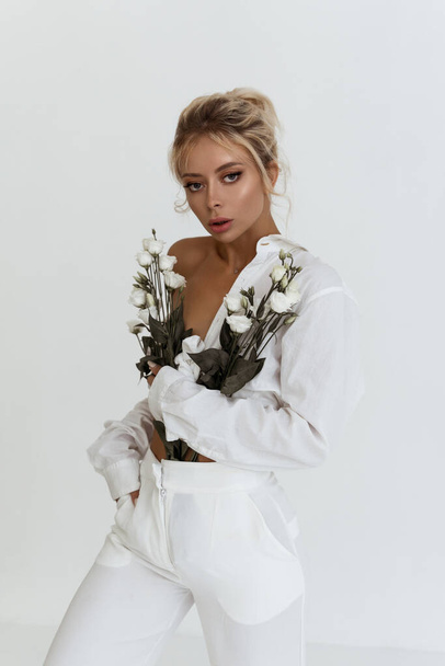 beautiful blonde model posing in white clothes. woman with updo hairstyle. natural make-up look. glamour lady holding flowers - Foto, Imagen