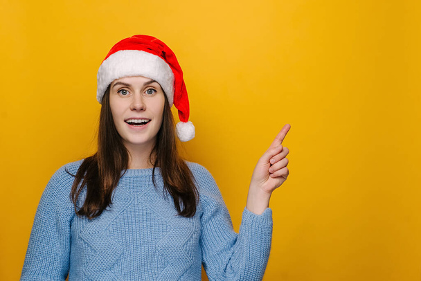 Emotional woman in Christmas hat, points fore finger aside on blank space, smiles happily, shows advertising, wears winter sweater, isolated over yellow wall. Happy New Year celebration merry holiday - Foto, imagen