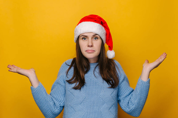 Doubting young girl shrug shoulders, looks uncertain, confused and looking to hand palms, wears Christmas hat and blue sweater, isolated on yellow studio wall. New Year celebration merry holiday - Foto, Imagem