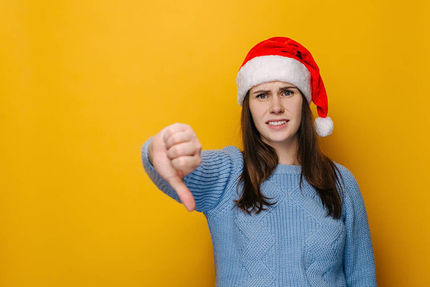 Discontent young woman shows disapproval sign, keeps thumb down, expresses dislike, frowns face in discontented, dressed in Christmas hat, isolated over yellow background. New Year holiday concept - Zdjęcie, obraz
