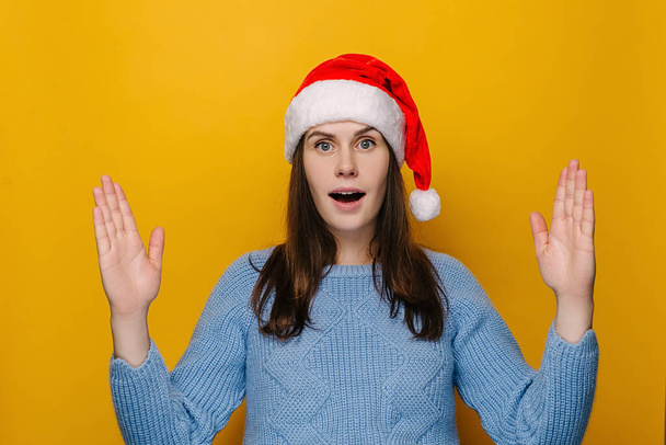 Σοκαρισμένη νεαρή γυναίκα σε χριστουγεννιάτικο καπέλο δείχνει μεγάλο μέγεθος χειρονομία με τα χέρια, ελκυστική κατάπληκτος κορίτσι, φοράει πουλόβερ, απομονώνονται σε κίτρινο φόντο στούντιο, εντυπωσιασμένος θηλυκό δείχνουν κάτι ευρύ - Φωτογραφία, εικόνα