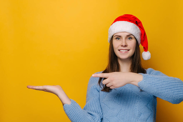 Positive young woman keeps palm raised, points with forefinger against yellow wall for advertisement, dressed in Christmas red hat and cozy blue sweater. Happy New Year celebration merry holiday - Fotoğraf, Görsel
