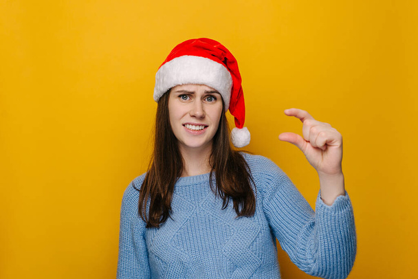 Young female shows very tiny object, shapes something small, wears Christmas hat and blue sweater, female looks in puzzlement, need some more, isolated over yellow studio wall. New Year concept - Foto, imagen