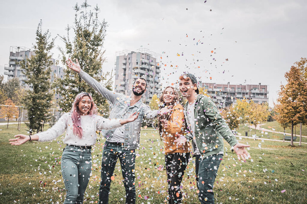 Grupo de amigos elegantes sonriendo y disfrutando de fiesta lanzando confeti colorido al cielo - chicos multiétnicos en el parque lanzando confeti colorido en el aire - Foto, Imagen