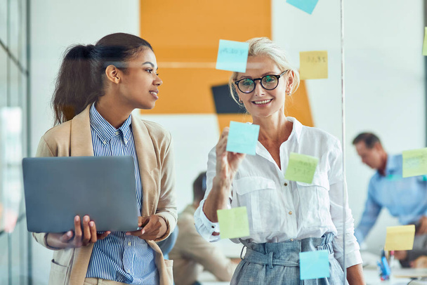 wo szczęśliwa kobieta współpracownicy wprowadzenie kolorowe lepkie notatki na szklanej ścianie i uśmiecha się stojąc w nowoczesnym biurze, ludzie biznesu pracujący razem - Zdjęcie, obraz