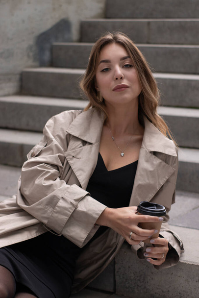 attractive brunette in fashionable trench and black dress sitting on grey concrete staircase with brown paper cup coffee - Фото, зображення