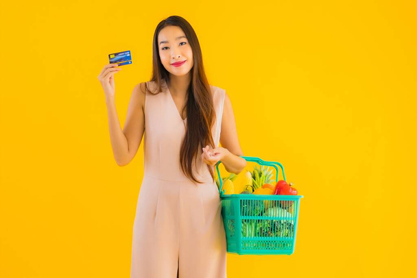 Портрет красива молода азіатка з продуктовим кошиком торгової сумки з супермаркету з кредитною карткою на жовтому ізольованому фоні
 - Фото, зображення