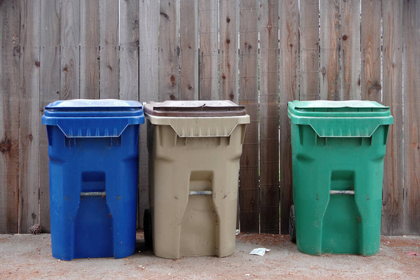 Três lixeiras para reciclagem, eliminação de lixo e resíduos de jardinagem, lado a lado em frente a uma grande parede de madeira - Foto, Imagem