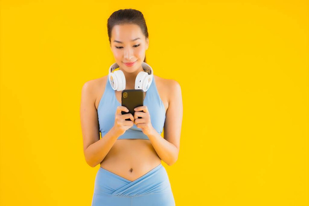 Portret piękna młoda azjatycka kobieta nosić strój sportowy ze smartfonem i słuchawką do słuchania muzyki na żółtym tle - Zdjęcie, obraz