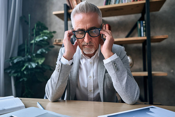 Tired adult businessman in glasses sitting at workplace in office and holding head on hands. Business concept - Fotografie, Obrázek