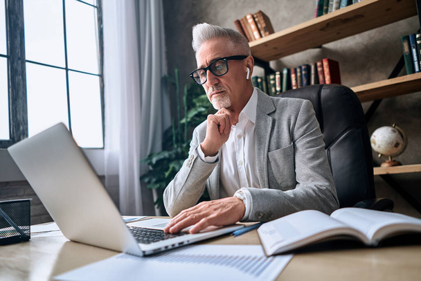 Mature serious man in glasses working online on computer while sitting in his office and holding a hand near the chin. Business concept - 写真・画像