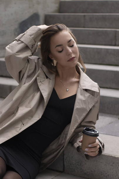 Çekici esmer, şık siperli ve siyah elbiseli gri beton merdivende kahverengi kağıt bardaklı kahve ile oturuyor. - Fotoğraf, Görsel