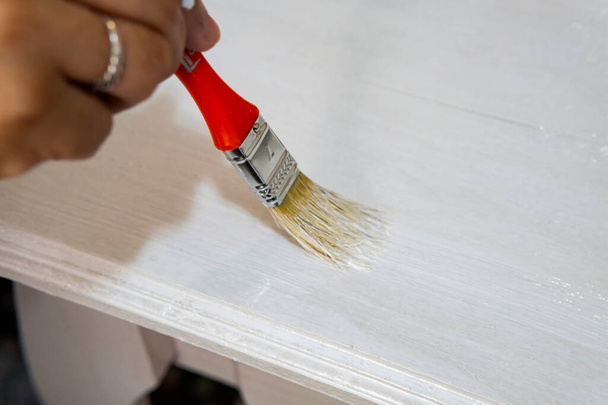 Szoros kilátás egy női kéz kezében egy kefét, és paiting fehér egy kis asztal. Fentről vették és lenéztek. Lőtt beltéri fehér fényes fény alatt - Fotó, kép