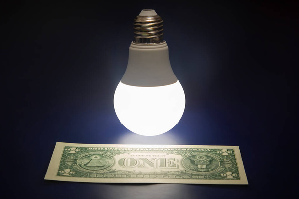 Lámpara led brillante y billete de un dólar sobre un fondo oscuro. Negocios y ahorros. Tecnología moderna y electricista - Foto, imagen