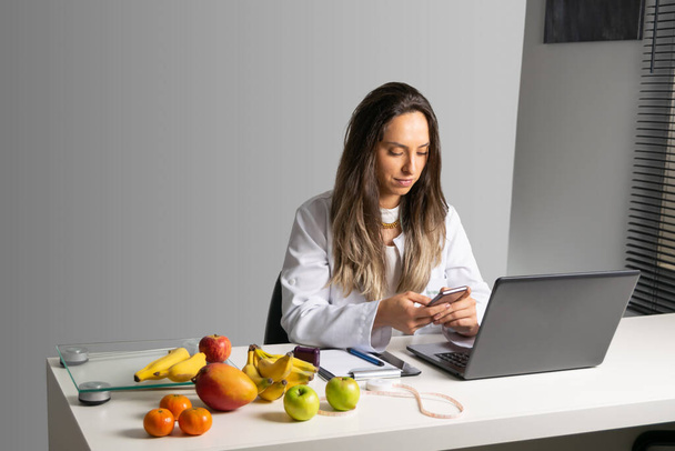 Χαμογελώντας γυναίκα διατροφολόγος στο γραφείο της, χρησιμοποιώντας κινητό τηλέφωνο, δείχνοντας υγιεινά λαχανικά και φρούτα. Υγιεινή και διατροφή. Τρόπος ζωής. - Φωτογραφία, εικόνα