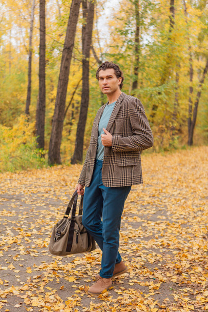 Tall handsome man dressed in a brown jacket walking with a bag on the autumn alley - Fotó, kép