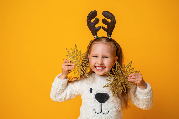 Happy little girl with deer antlers with Christmas stars on yellow background. child is happy, having fun and preparing for Christmas. Advertising. Copy space. - Fotoğraf, Görsel