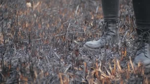 Shot from below in slow motion of persons legs in leather stylish youth boots standing on the burnt grass after serious fire in field. Man stomps on burned earth, ashes fly up in air from the wind. - Footage, Video
