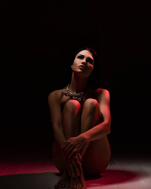 Sensual naked romantic woman in jewelry necklace sits on the floor with her arms around her knees and looks up - 写真・画像