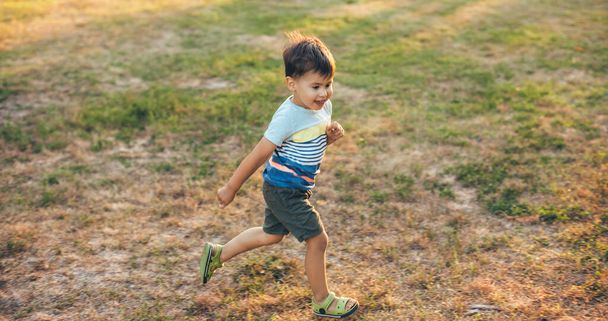 Correr caucasiano menino está sorrindo em um campo verde em um parque durante uma caminhada de verão - Foto, Imagem