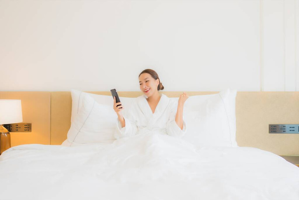 肖像画美しい若いアジアの女性は寝室のインテリアでベッドの上にスマートフォンを使用します - 写真・画像