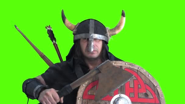 Viking krijger een boer in helm met hoorns en schild, gewapend met een bijl voor de strijd, slaat agressief geschilderd houten schild met grote bijl. volwassen man op groene achtergrond - Video