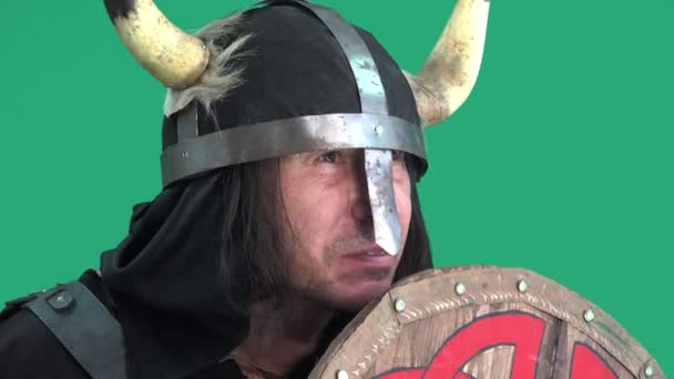 Mature man in helmet with horns. Aggressive viking warrior scares enemies by biting with crooked teeth wooden shield. Portrait in green background - Footage, Video