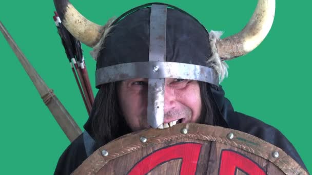Agresivo guerrero vikingo asusta a los enemigos al morder con los dientes torcidos escudo de madera. Hombre maduro en casco con cuernos. Retrato en fondo verde - Metraje, vídeo