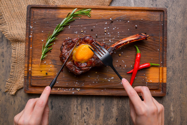 Barbecue tomahawk steak with spices served on a rustic wooden board over wooden background. BBQ concept, grilled meat steak, autumn seasonal dish. Eating steak with knife and fork. - Foto, imagen