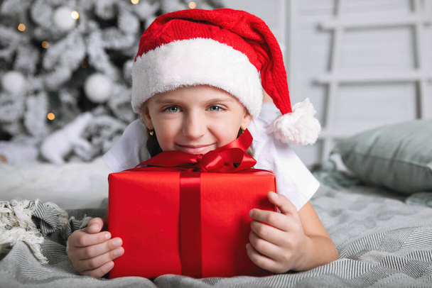 Dziewczyna w czerwonej czapce z prezentem świątecznym. Leży na łóżku, przytula pudełko na tle choinki. - Zdjęcie, obraz