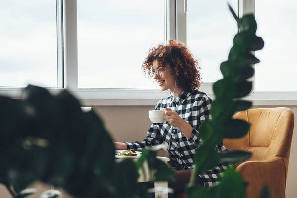 Smiling young businesswoman with curly hair and glasses is drinking a cup of tea while eating something at the laptop - Foto, Imagen