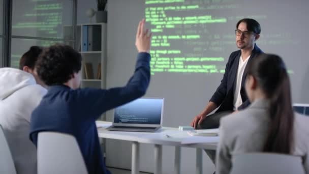 Середній знімок ІТ-репетитора пояснює матеріал уроку про програму кодування в сучасному класі з екранним проектором на фоні, і один зі студентів піднімає руку, щоб поставити питання
 - Кадри, відео