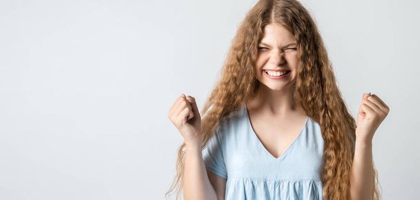 Life is wonderful. Joyful girl with curly long hair keeps fists clenched and exclaims in triumph, celebrates success, isolated over white background with blank space for your advertisement - Photo, image
