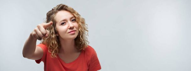 Pretty Young woman with curly hair in casual t shirt looking at the camera and pointing her finger at you. Studio shot, white background. Human emotions, facial expression concept - Photo, Image