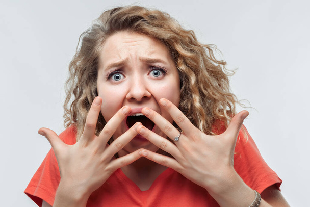 I'm afraid. Image of scared blonde girl with curly hair in casual t shirt covering her mouth with hands. Fright, phobia, panic attack, horror and facial expression concept - Photo, Image