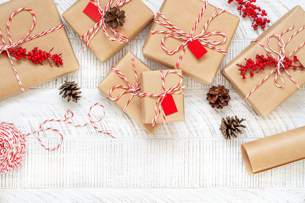 Preparing christmas gifts with craft paper, striped twine string, branches with red berries and pine cones on white wooden background. New year flat lay. Top view of decorated boxes. Copy space. - Foto, Imagem