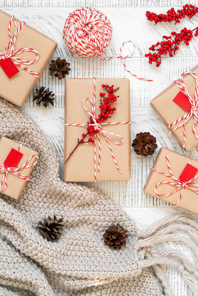 Handcrafted gifts. Christmas flat lay with pine cones, presents wrapped in craft paper and decorated with striped baker's twine string and holly berries on wooden background. Top view. - Photo, image