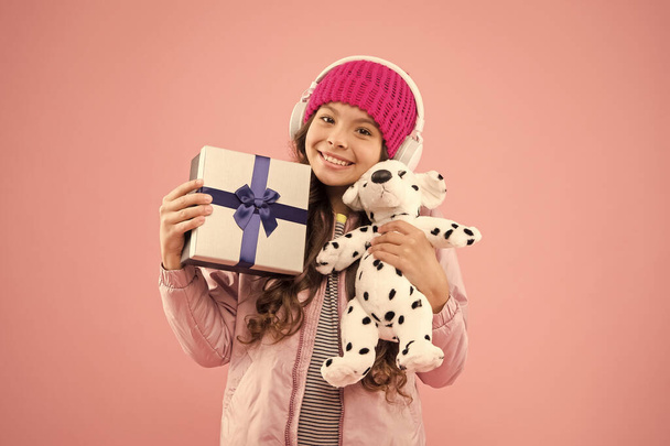 Girl little fashionable cutie with headphones carry soft toy. Take favorite toy with you. Happy childhood concept. Best gifts for teen girls. Stylish schoolgirl play with dog toy and hold gift box - Photo, image