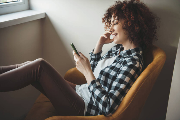 Кудрявая кавказка в повседневной одежде расслабляется в кресле, разговаривая по телефону у окна - Фото, изображение