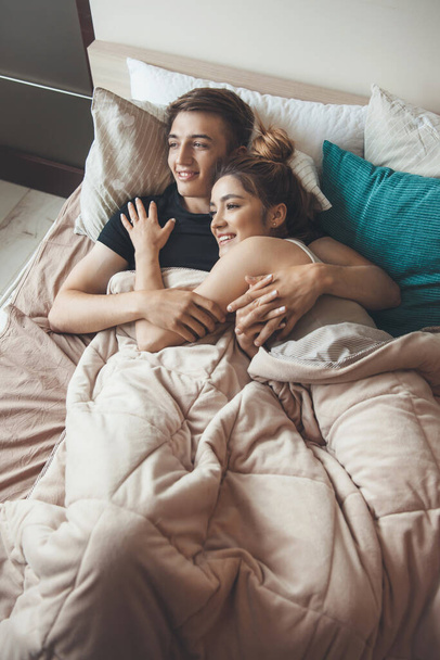 Вид зверху фото кавказької пари лежить на ліжку, обіймаючись під ковдрою і посміхаючись вранці
 - Фото, зображення
