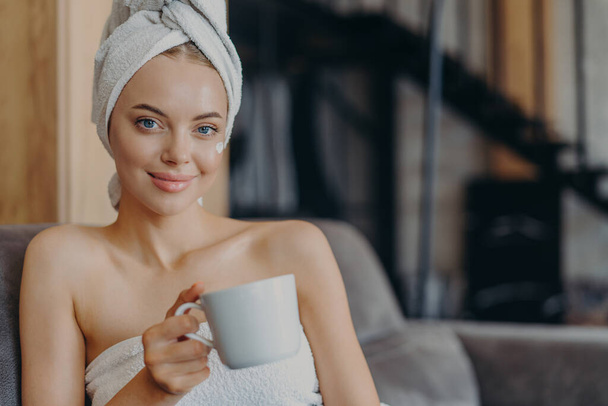 Adorable woman with natural makeup and healthy skin, poses bare shoulders, wrapped in bath towel, drinks tea or coffee, sits on comfortable sofa against home interior. People, hygiene, relaxation - Photo, Image