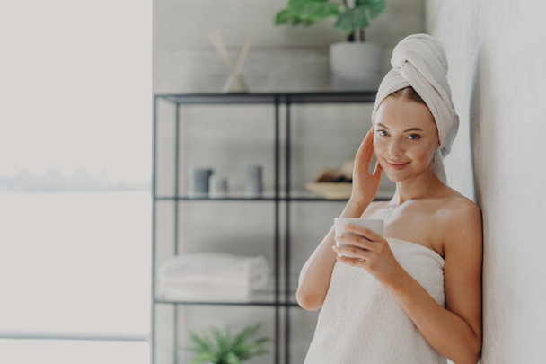 Pleased attractive woman has natural beauty, glowing smooth healthy skin, touches face, enjoys spa procedures, wears white bath towel on head and around body, drinks tea, poses in cozy bathroom - Photo, Image