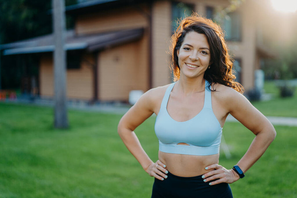 Beautiful pleased curly haired woman in good shape, keeps hands on waist, dressed in cropped top, smiles gladfully, has sporty body, poses outdoor against house background. Morning workout concept - Photo, Image