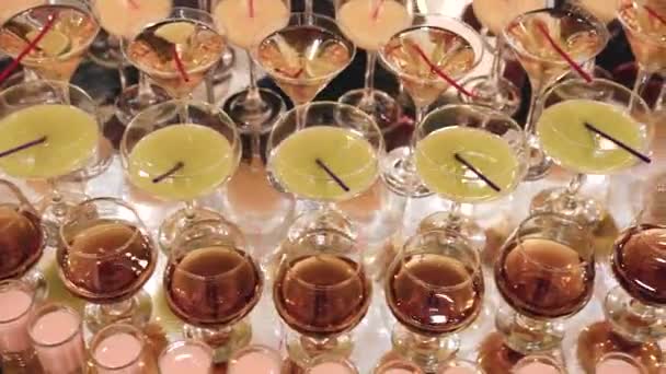 Bij een duur buffet staan heldere glazen van verschillende vormen en maten gevuld met kleurrijke elegante alcoholische dranken op een spiegeloppervlak dicht bij elkaar. Snelle camerabeweging - Video
