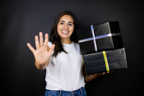 Mladá krásná žena drží dárky na izolovaném černém pozadí ukazuje a ukazuje s prsty číslo pět a zároveň se usmívá sebevědomě a šťastně - Fotografie, Obrázek