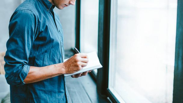 man making notes in a notebook, standing near the window. - Φωτογραφία, εικόνα