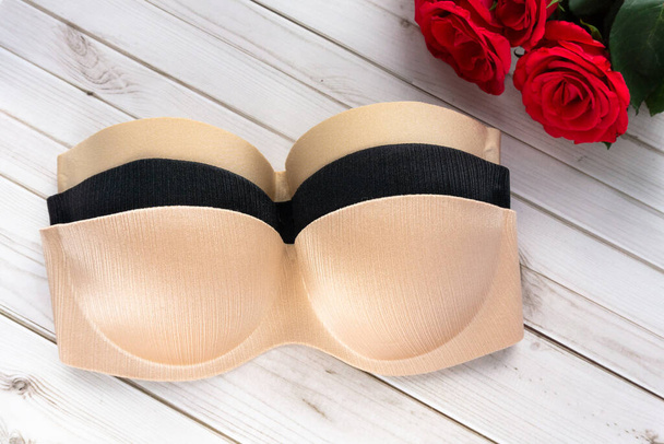 beige and black strapless nude bra on a wooden texture background next to rose flowers . Copy space for text input on health care and fitness. Balconette bra and lingerie for woman, top view. - Photo, Image