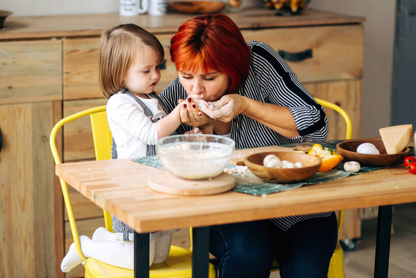 Kochen Sie gemeinsam zu Hause. Rothaarige Großmutter und kleine Enkelin kochen in einem gemütlichen Haus Pizza. Viel Spaß in der Küche - Foto, Bild