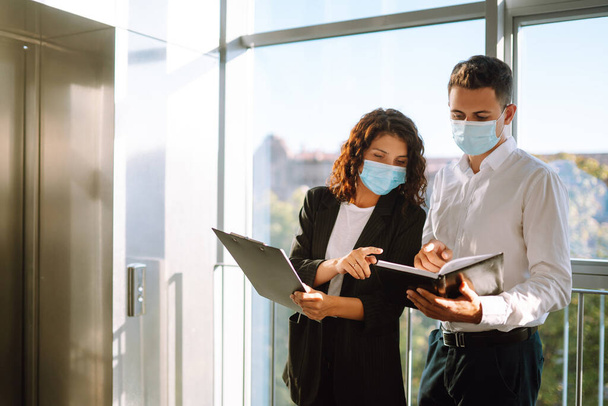 Two colleagues in protective face masks in modern office discussing together work issues standing near elevator. Business People back at work in office after quarantine. Covid-19. - Fotoğraf, Görsel